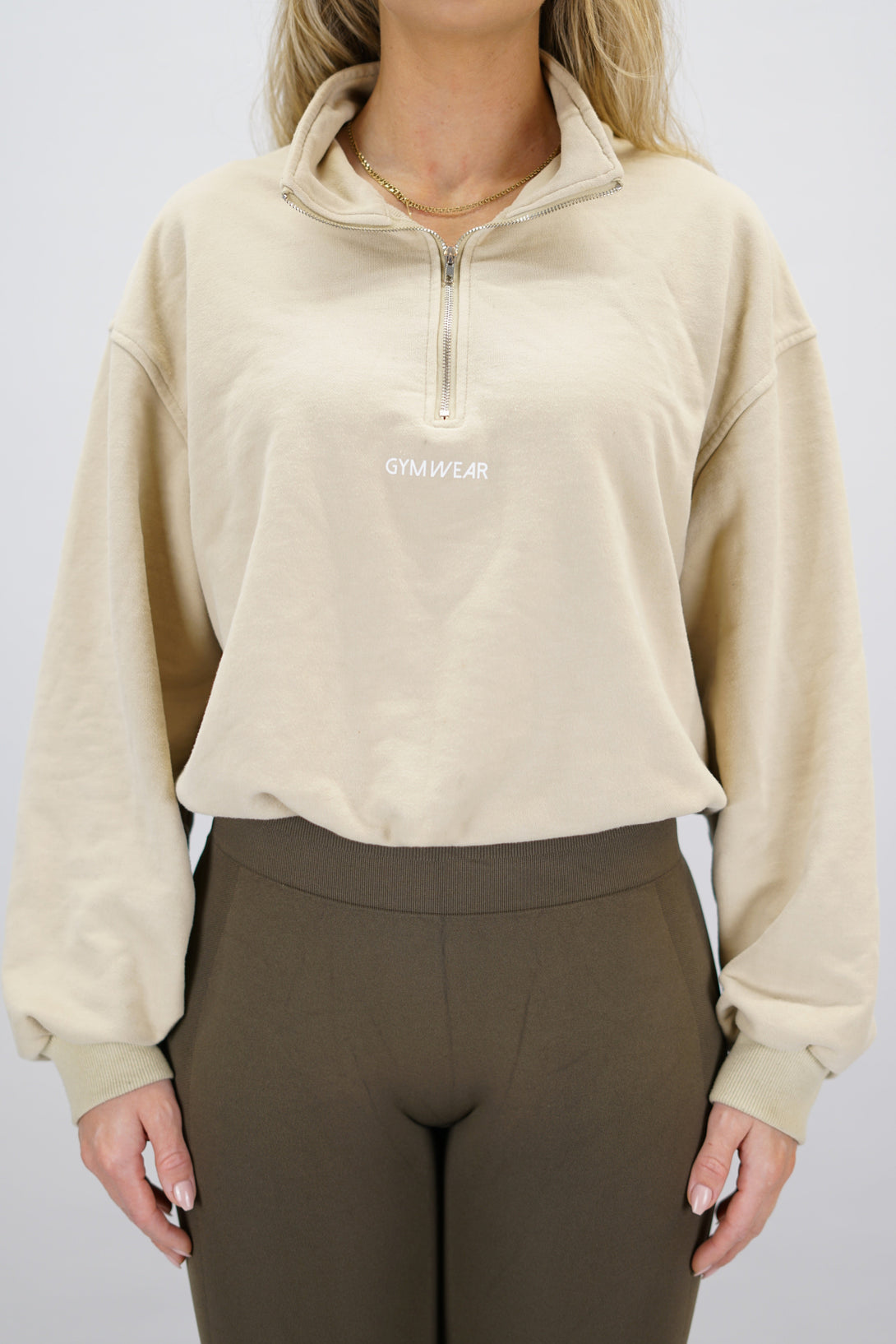 Modell bär en beige Ayla Half-Zip Long Sleeve, en långärmad träningströja med halvdragkedja vid halsen och liten Gymwear-logotyp centrerat på bröstet och symbolen g i nacken