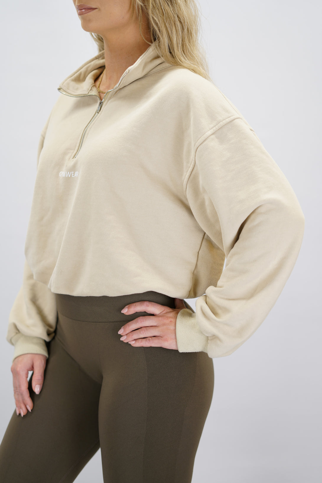 Modell bär en beige Ayla Half-Zip Long Sleeve, en långärmad träningströja med halvdragkedja vid halsen och liten Gymwear-logotyp centrerat på bröstet och symbolen g i nacken