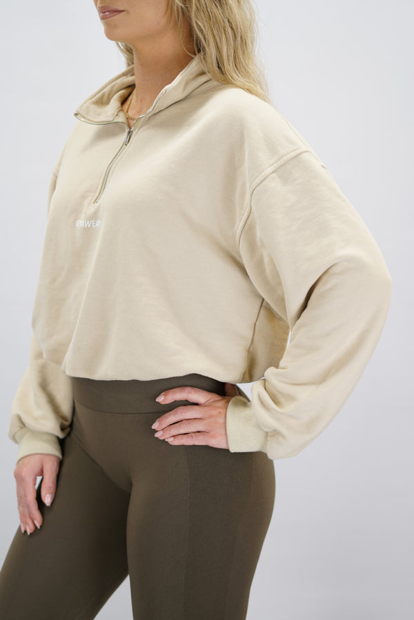 Modell bär en beige Ayla Half-Zip Long Sleeve, en långärmad träningströja med halvdragkedja vid halsen och liten Gymwear-logotyp centrerat på bröstet och symbolen g i nacken