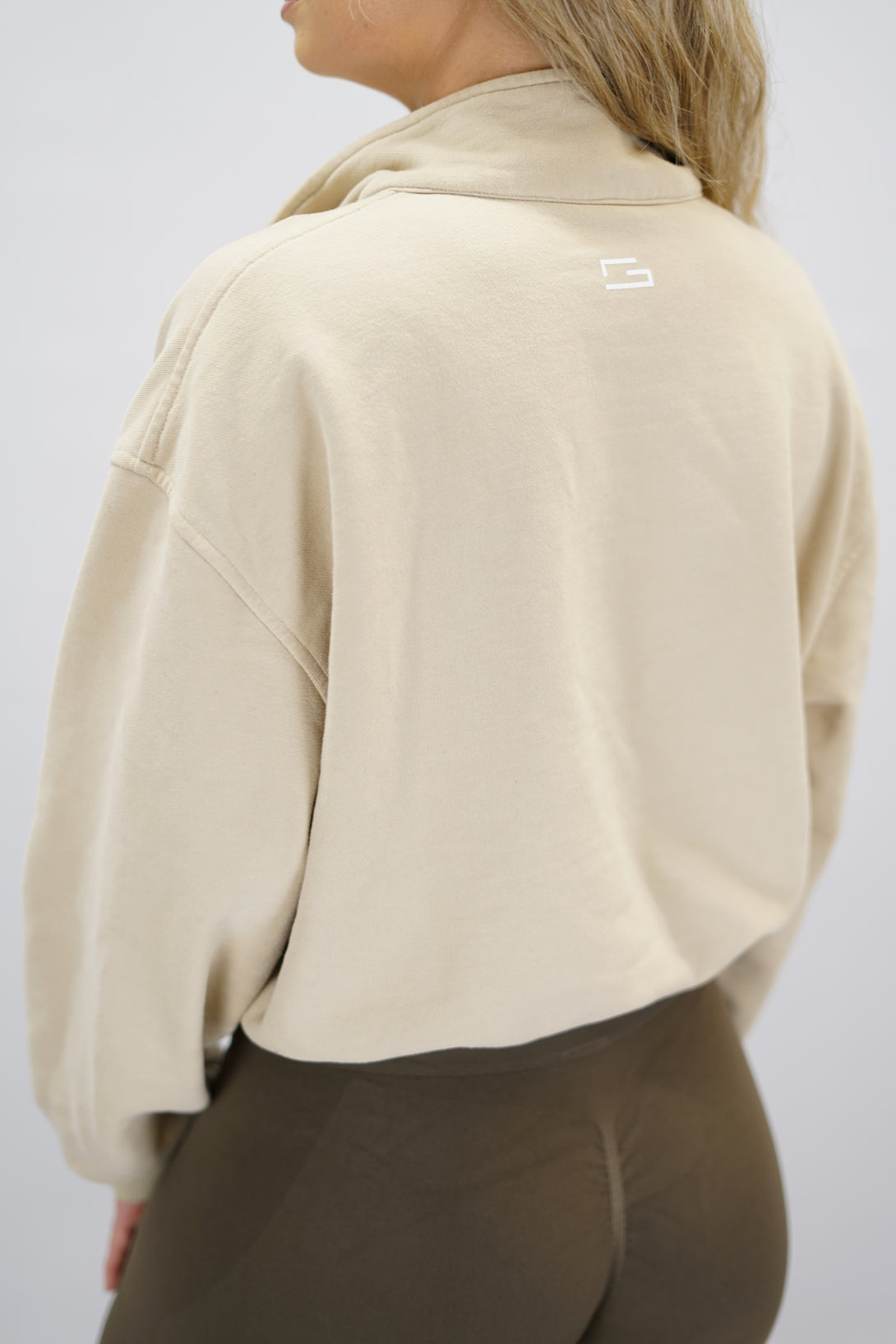 Modell bär en beige Ayla Half-Zip Long Sleeve, en långärmad träningströja med halvdragkedja vid halsen och liten Gymwear-logotyp centrerat på bröstet och symbolen g i nacken