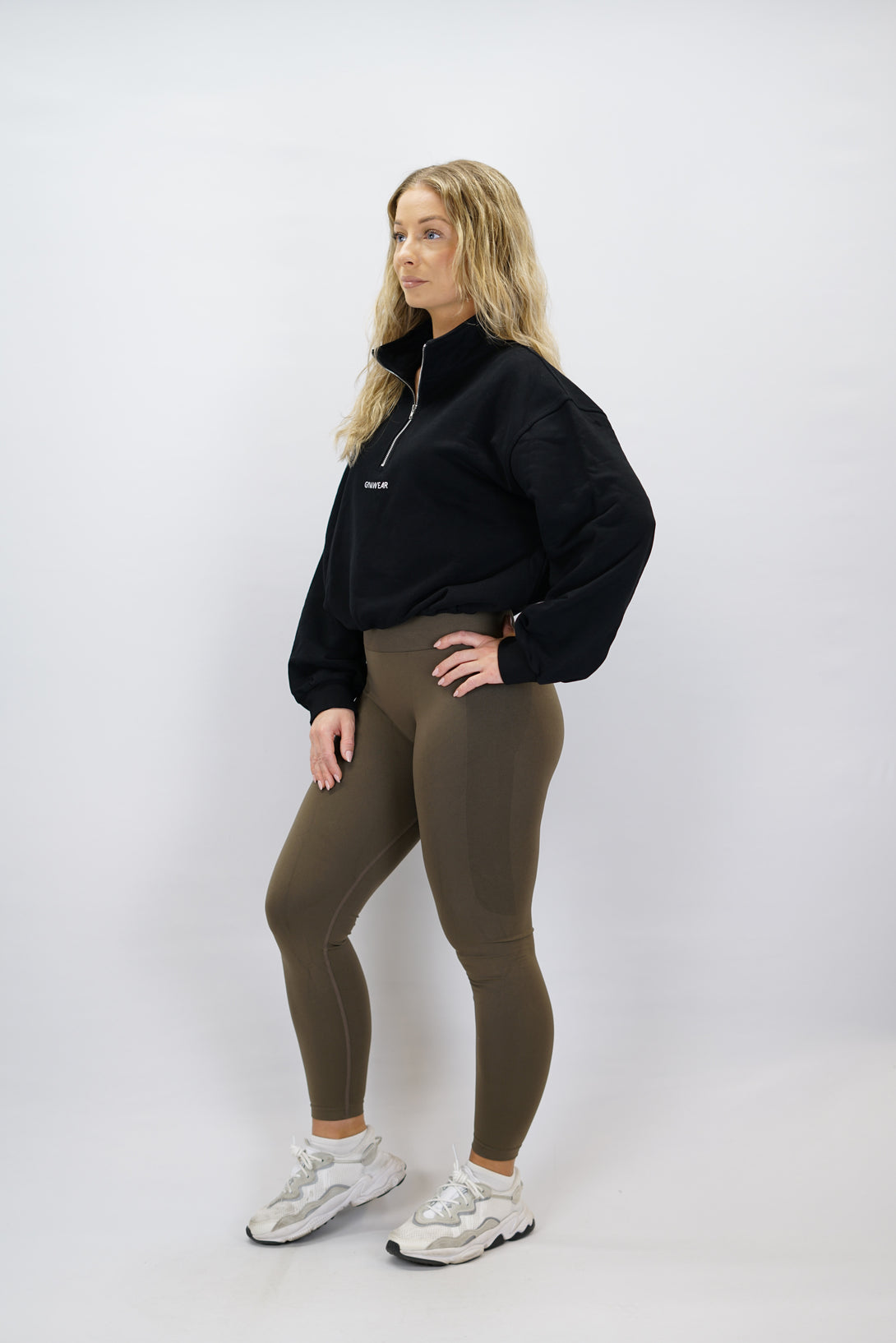 Modell bär en svart Ayla Half-Zip Long Sleeve, en långärmad träningströja med halvdragkedja vid halsen och liten Gymwear-logotyp centrerat på bröstet och symbolen g i nacken.