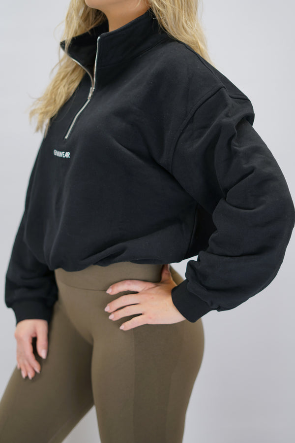 Modell bär en svart Ayla Half-Zip Long Sleeve, en långärmad träningströja med halvdragkedja vid halsen och liten Gymwear-logotyp centrerat på bröstet, och symbolen g i nacken.
