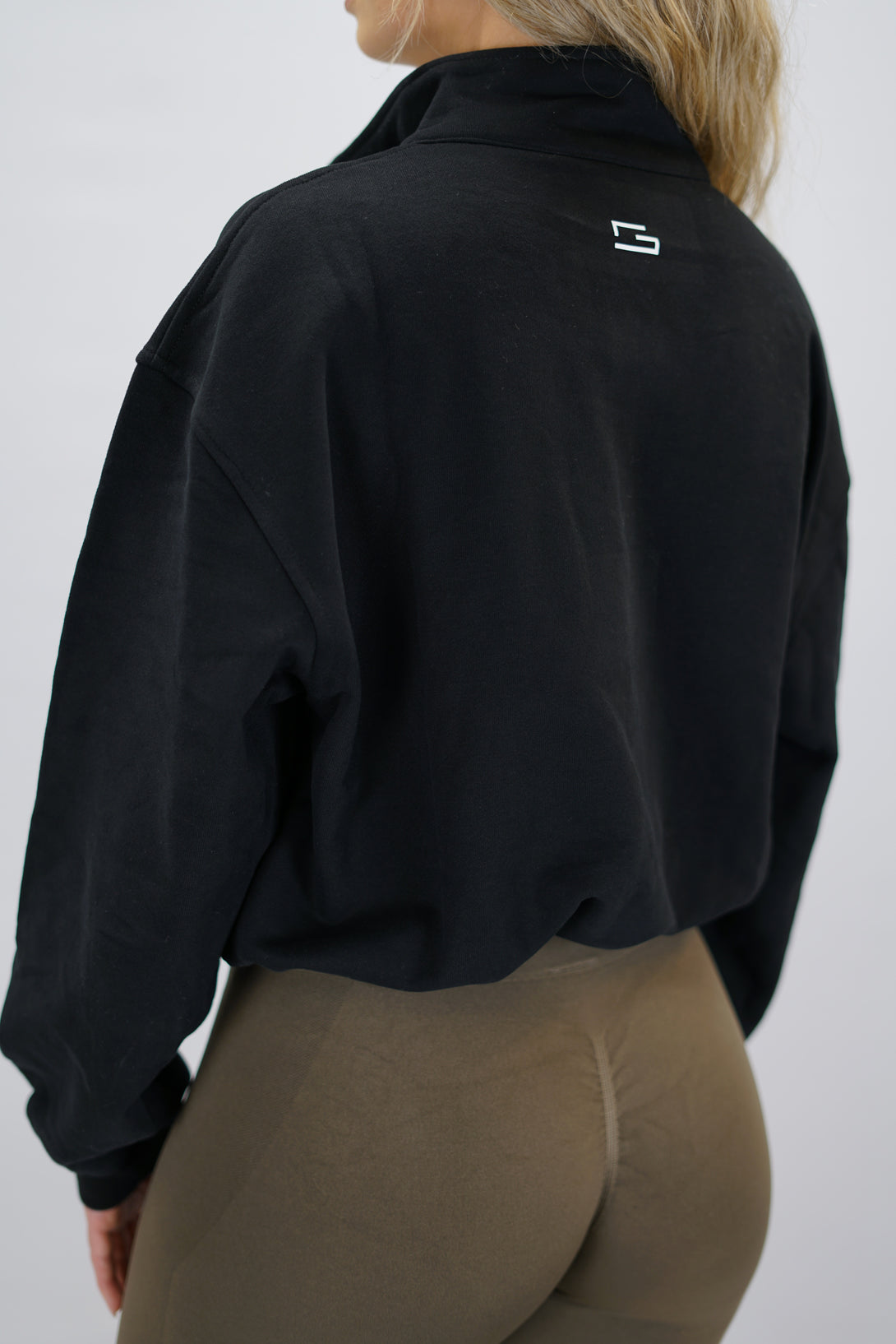 Modell bär en svart Ayla Half-Zip Long Sleeve, en långärmad träningströja med halvdragkedja vid halsen och liten Gymwear-logotyp centrerat på bröstet och symbolen g i nacken