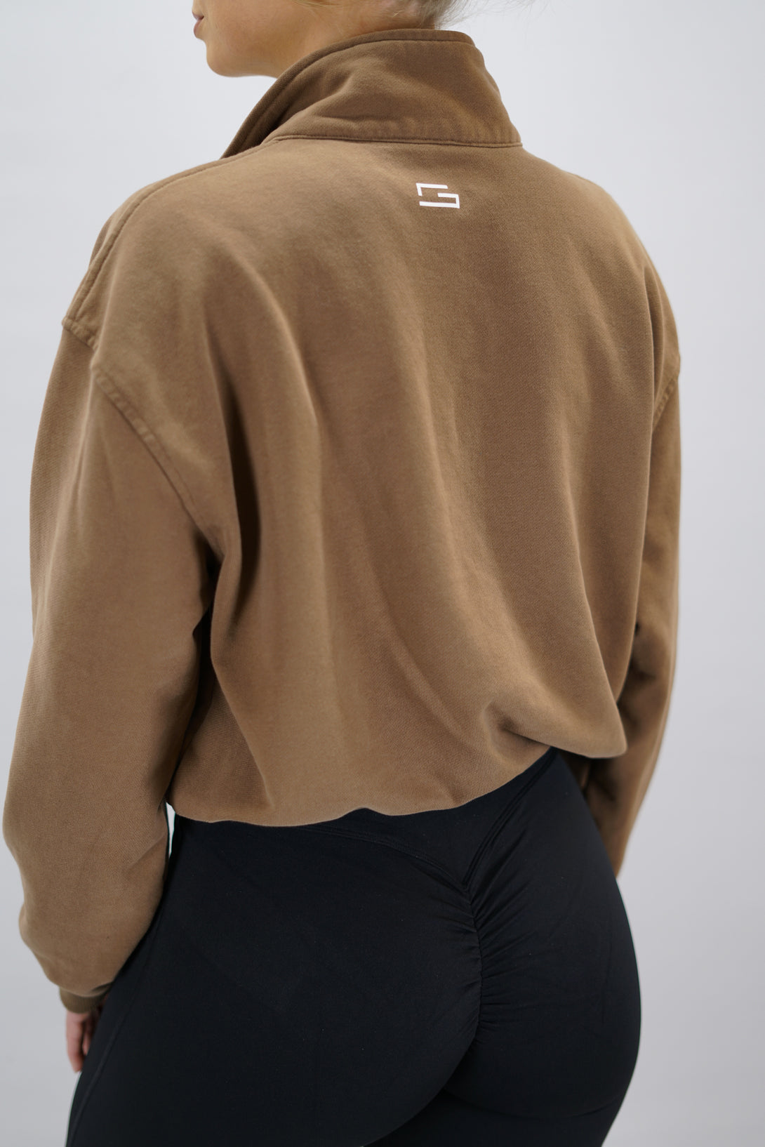 Modell bär en brun Ayla Half-Zip Long Sleeve, en långärmad träningströja med halvdragkedja vid halsen och liten Gymwear-logotyp centrerat på bröstet och symbolen g i nacken 