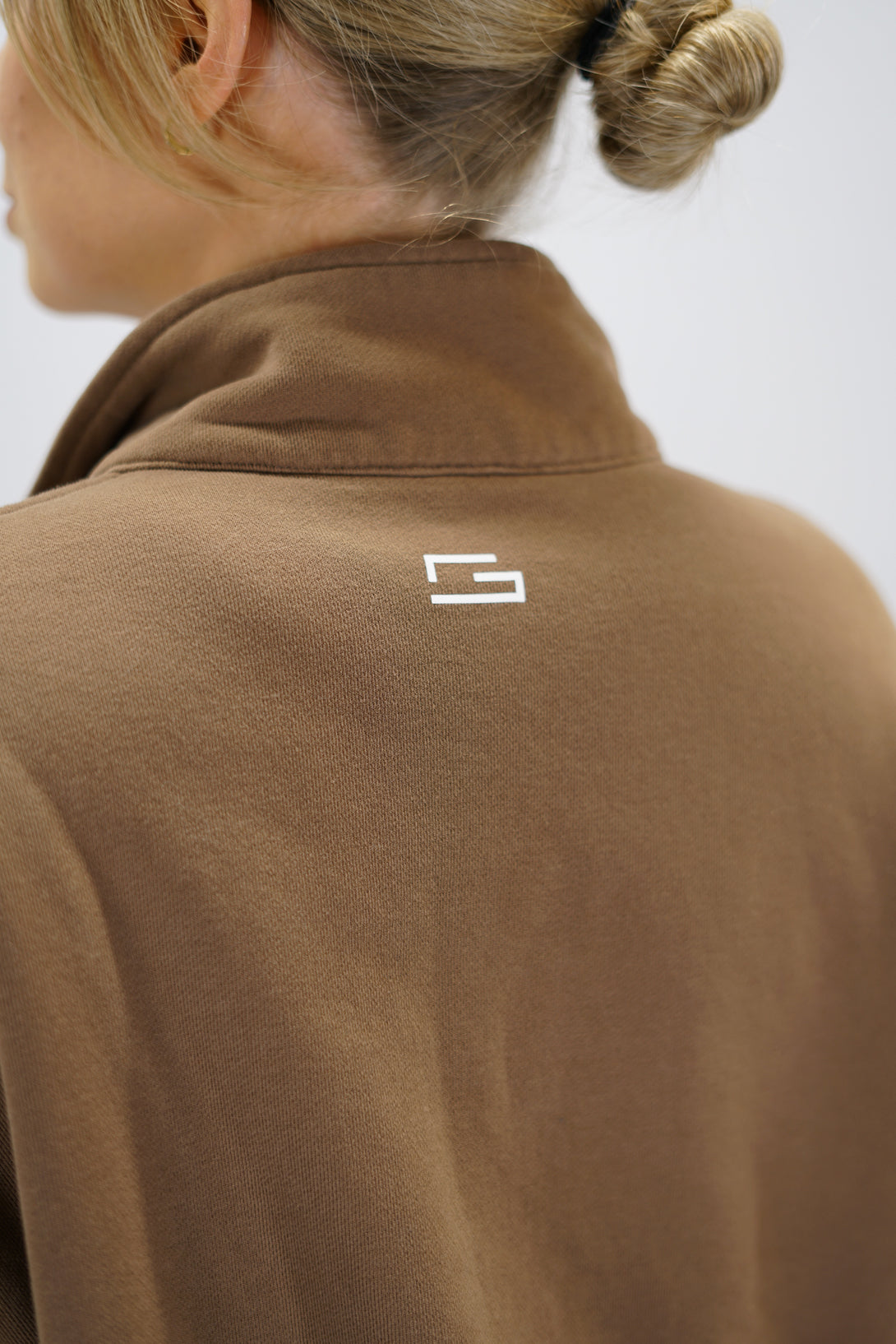 Modell bär en brun Ayla Half-Zip Long Sleeve, en långärmad träningströja med halvdragkedja vid halsen och liten Gymwear-logotyp centrerat på bröstet och symbolen g i nacken 