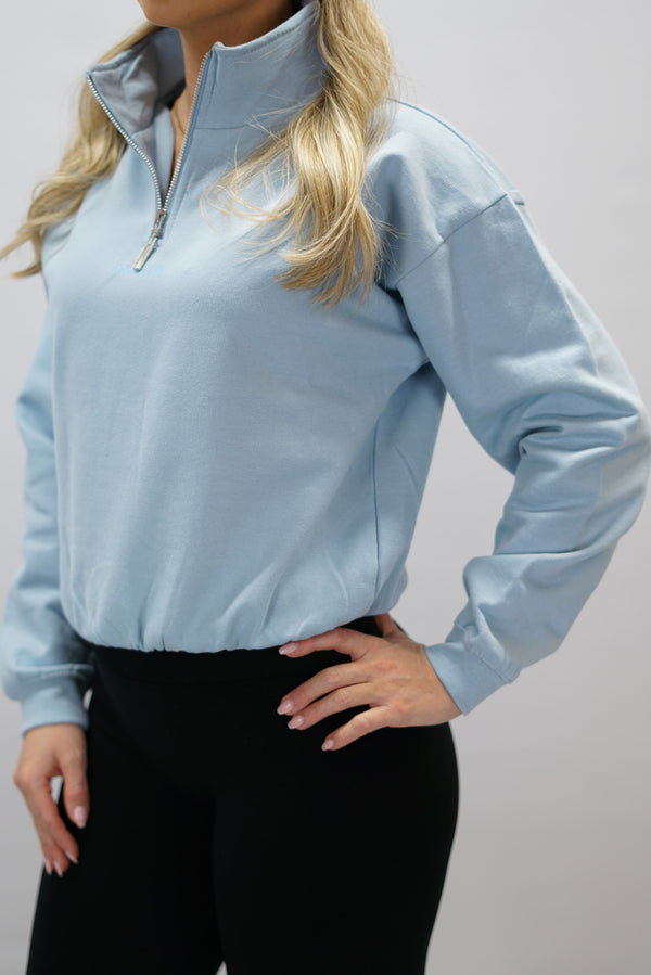 Blå Delia Half-Zip Long Sleeve, en långärmad tröja i 100% bomull med halvdragkedja vid halsen och diskret ton-i-ton Gymwear-logotyp centrerat på bröstet.
