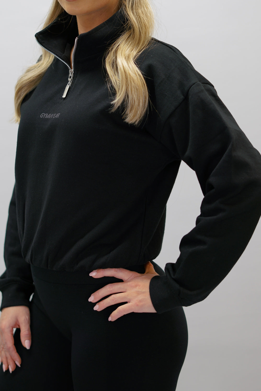 Svart Delia Half-Zip Long Sleeve, en långärmad tröja i 100% bomull med halvdragkedja vid halsen och diskret ton-i-ton Gymwear-logotyp centrerat på bröstet.