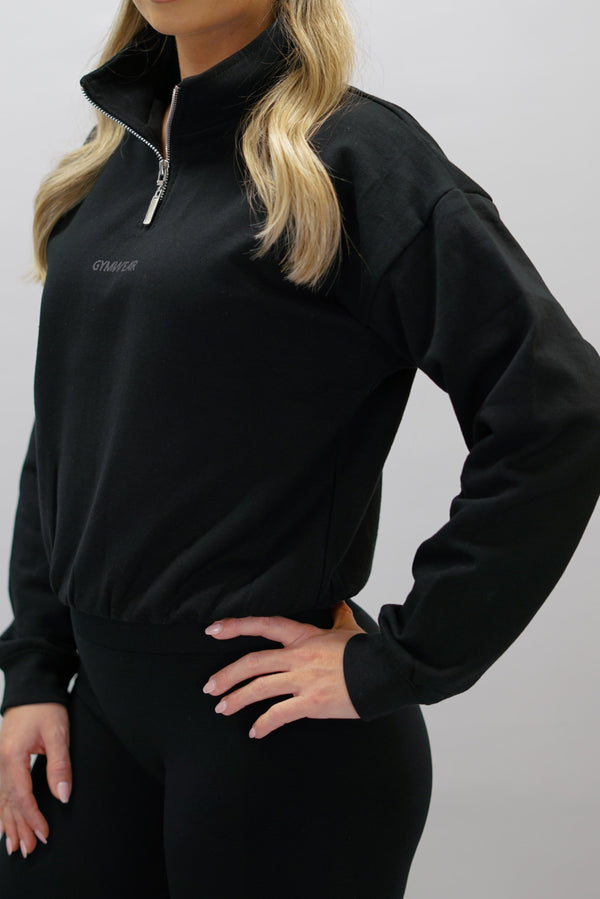 Svart Delia Half-Zip Long Sleeve, en långärmad tröja i 100% bomull med halvdragkedja vid halsen och diskret ton-i-ton Gymwear-logotyp centrerat på bröstet.