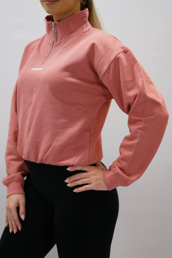 Gammaldags rosa Delia Half-Zip Long Sleeve, en långärmad tröja i 100% bomull med halvdragkedja vid halsen och diskret ton-i-ton Gymwear-logotyp centrerat på bröstet.