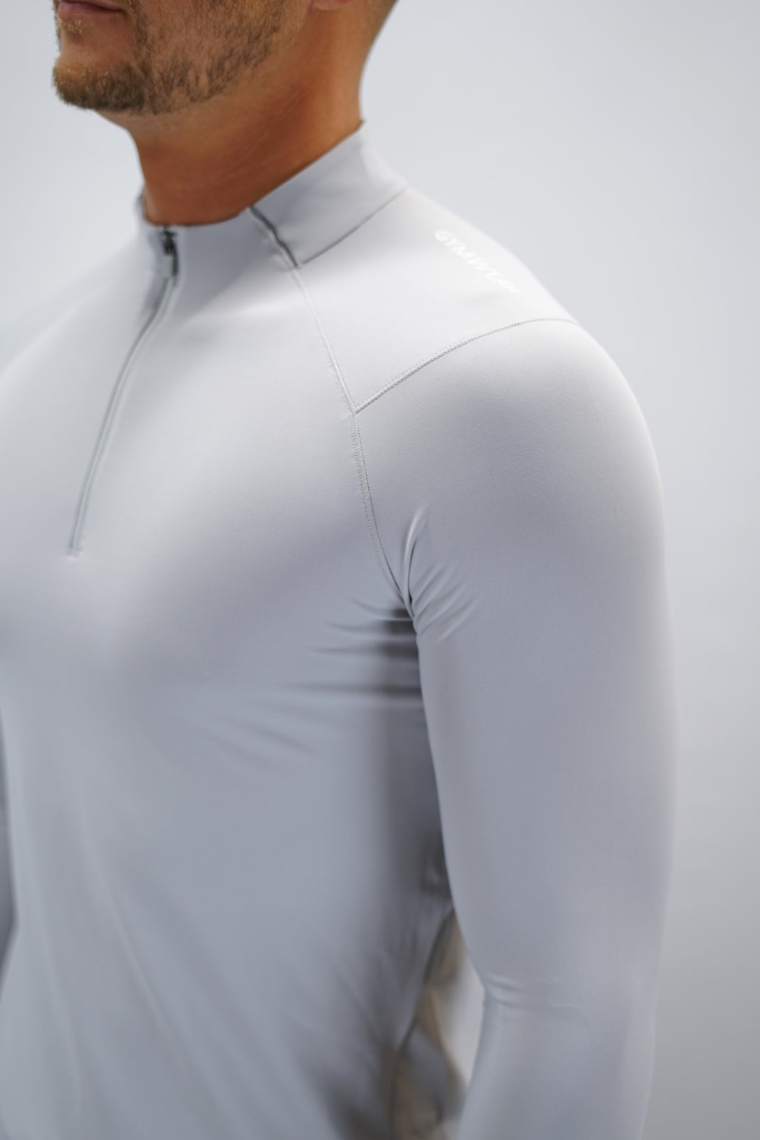 Ljusgrå Zayne Half-Zip Long Sleeve – Reflective Detail - långärmad träningströja med halvdragkedja, tumhål, klockutrymme, reflekterande detalj vid kragen och Gymwear-logga på framsidan