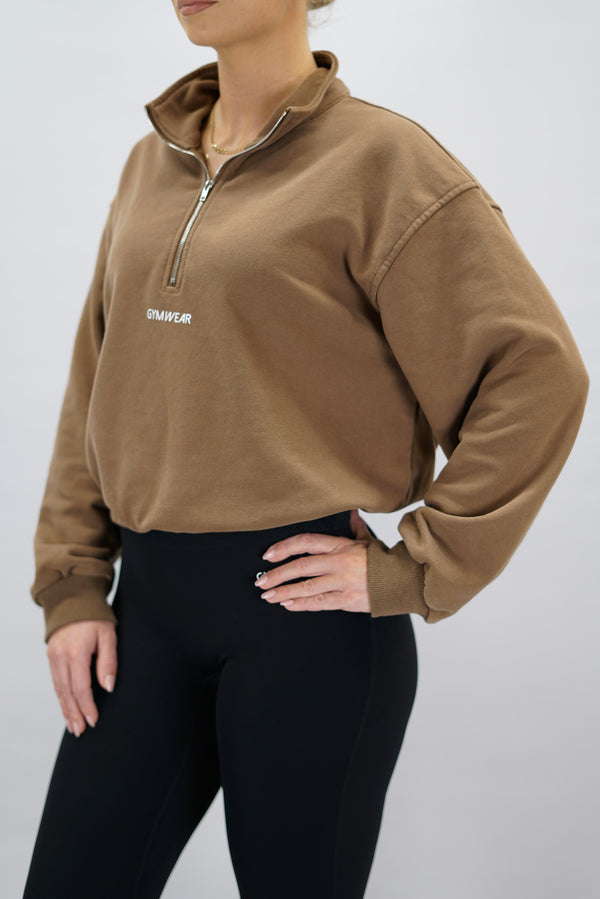 Modell bär en brun Ayla Half-Zip Long Sleeve, en långärmad träningströja med halvdragkedja vid halsen och liten Gymwear-logotyp centrerat på bröstet och symbolen g i nacken 
